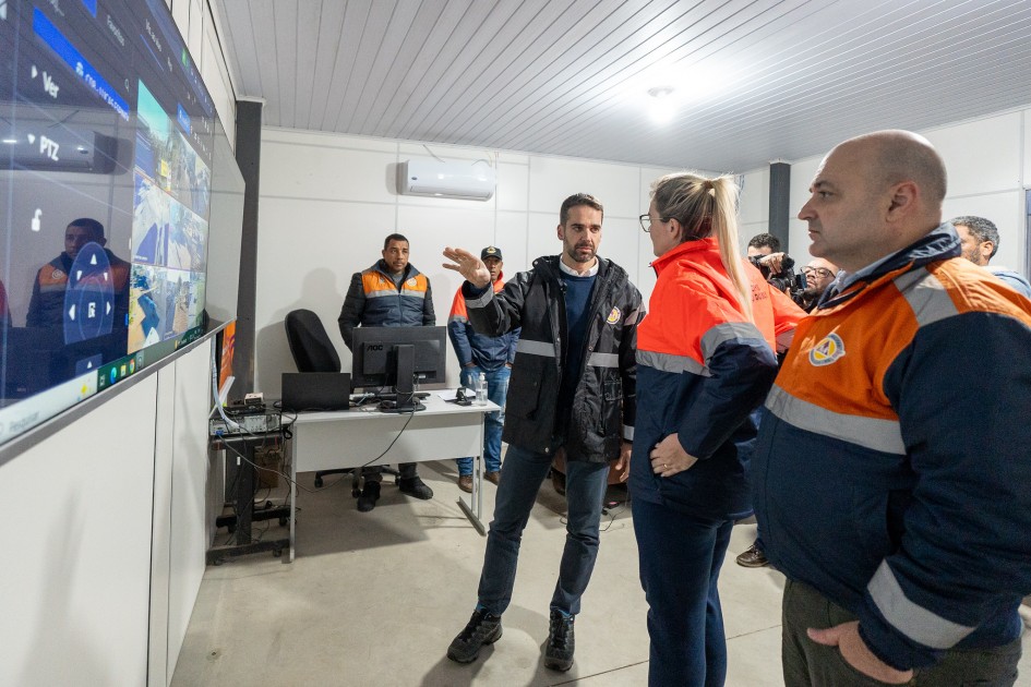 Foto mostra uma sala de controle, onde várias pessoas, incluindo Leite, usam uniformes com jaquetas laranja e azul e estão focadas em monitores. Leite aponta para uma grande tela na parede que exibe várias câmeras e opções de controle.