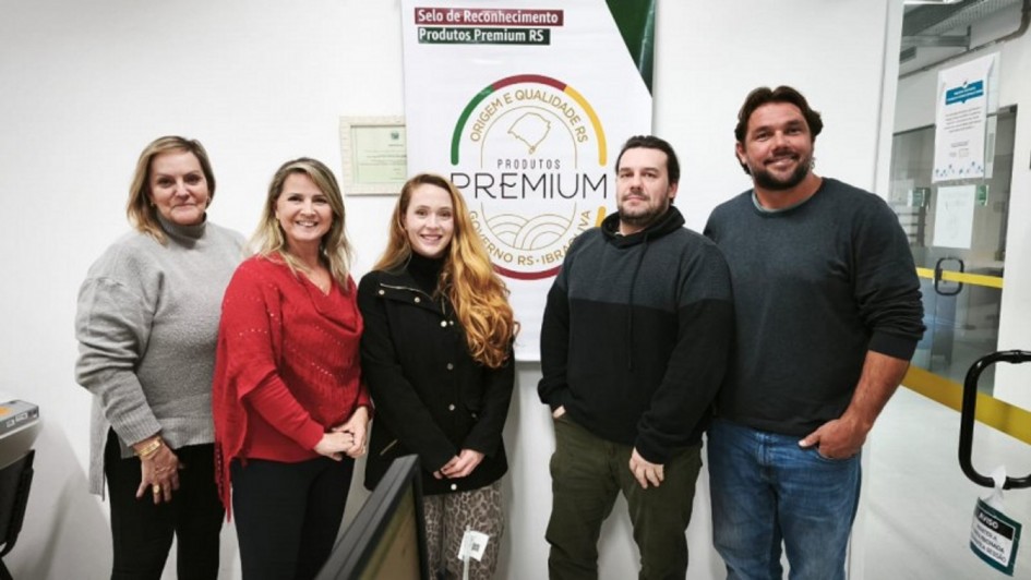 A foto mostra um grupo de cinco pessoas, três mulheres e dois homens, posando em pé na frente de um painel promocional do Programa Produtos Premium. O painel branco tem um logotipo no topo que diz "Selo de Reconhecimento Produtos Premium RS", com um ícone de "ORIGEM E QUALIDADE RS" e a palavra "PREMIUM" em destaque. À esquerda, as duas mulheres sorriem. A mulher do centro tem cabelos ruivos e usa uma jaqueta preta. À direita, os dois homens sorriem. O ambiente parece ser um corredor ou recepção, com paredes claras e portas no fundo. A cena sugere a concessão de um selo de qualidade para produtos gaúchos.