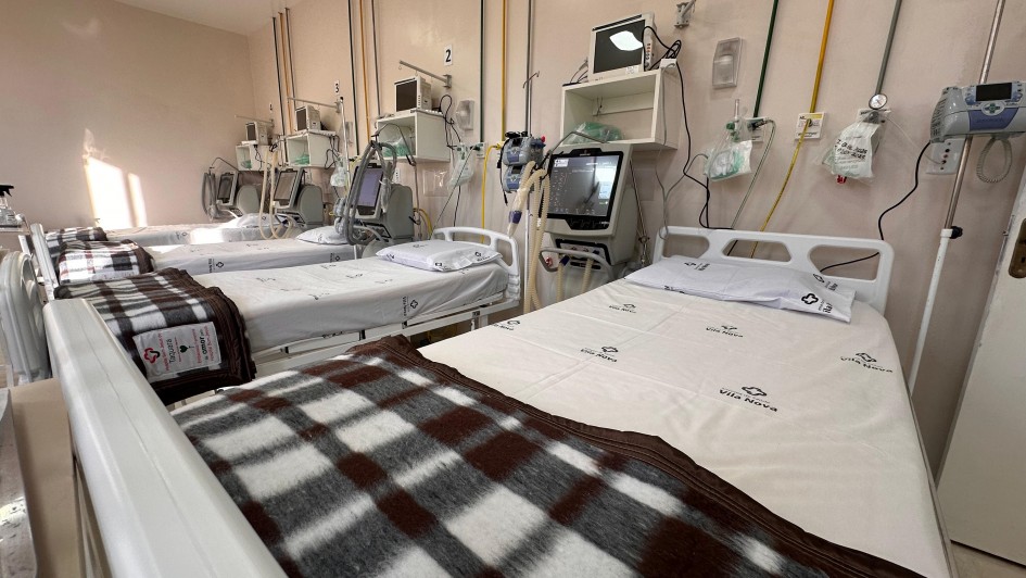 A imagem mostra um quarto hospitalar com vários leitos organizados em fila. Cada cama está equipada com lençóis brancos e cobertores xadrez em tons de marrom. Também há equipamentos médicos, como monitores, suportes de soro e outros dispositivos hospitalares.