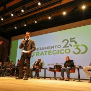 A foto mostra o governador Eduardo Leite de pé no palco, centralizado, discursando. Ele é um homem branco, vestindo um terno escuro, camisa branca e gravata preta. Leite segura um microfone com a mão esquerda e um papel com a direita, olhando para a plateia (não visível). Atrás dele, um grande telão branco exibe a frase "PLANEJAMENTO ESTRATÉGICO" e os números "25 30" em destaque. À esquerda do palco, um músico de cabelos crespos toca um violoncelo vermelho, com outros músicos parcialmente visíveis ao fundo, sugerindo uma orquestra. À direita de Leite, três homens adultos estão sentados em poltronas escuras. O palco é de madeira clara e o fundo apresenta cortinas escuras.