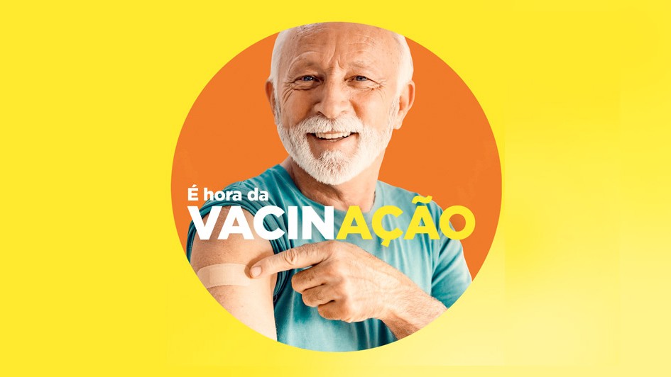 Card em fundo amarelo com um círculo em cor laranja ao centro. No círculo, há uma pessoa idosa de cabelo e barba branca, com camiseta azul. Ela aponta para um curativo no braço. A imagem traz o texto: “É hora da VACINAÇÃO”.