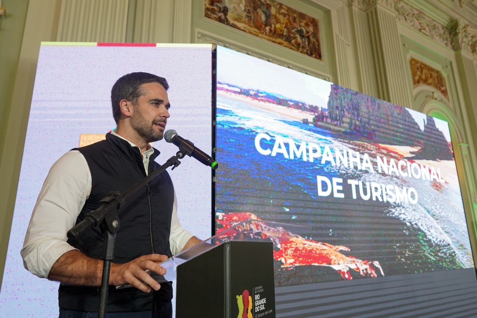 A imagem mostra o governador Eduardo Leite discursando em um evento. Ele está em pé, vestindo uma camisa branca e um colete escuro, falando em um microfone. À direita dele, há uma grande tela exibindo a frase "CAMPANHA NACIONAL DE TURISMO" sobre uma imagem de paisagem, que parece incluir uma praia e rochas, e uma imagem abstrata em tons de vermelho e laranja na parte inferior. Ao fundo, acima da tela, pode-se ver parte de uma parede com afrescos ou pinturas clássicas.