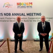 
A imagem mostra duas pessoas, aparentemente homens, de pé e sorrindo para a câmera em frente a um grande banner. Ambos estão usando ternos escuros. O banner, ao fundo, é predominantemente branco e exibe o logotipo do "New Development Bank" com a inscrição "10 years" (10 anos), e um cabeçalho vermelho com "NDB2025 - 10TH ANNUAL MEETING - RIO DE JANEIRO, BRAZIL - JULY 4-5, 2025". Abaixo, em letras grandes, está escrito "2025 NDB ANNUAL MEETING", seguido de um texto menor que parece ser o tema do evento: "Development: Fostering Innovation, Cooperation, and Impact through a Multilateral Development Bank for the Global South". Há um pequeno ícone de painéis solares no canto inferior direito do banner. Duas cordas de isolamento douradas com suportes escuros marcam o espaço à frente das pessoas.