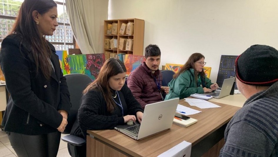 A foto mostra quatro pessoas em um ambiente interno, que parece ser um escritório ou sala de atendimento. À esquerda, uma mulher de cabelos castanhos escuros e blazer preto está de pé, olhando para a frente. No centro, sentada em uma mesa de madeira clara, uma mulher de cabelos castanhos e suéter preto digita em um notebook. Ao seu lado, um homem de cabelo escuro e jaqueta vinho também está sentado, olhando para o lado. Mais à direita, outra mulher de óculos e jaqueta verde está sentada, digitando em outro laptop. No canto inferior direito, parte da cabeça de um homem de gorro preto é visível, de costas para a câmera. Ao fundo, há quadros coloridos e prateleiras com caixas de papelão. A cena sugere um mutirão ou atendimento para regularização de documentos.