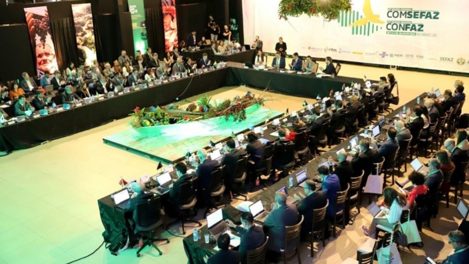 Foto mostra uma grande reunião em formato de mesa em U, com dezenas de pessoas sentadas ao redor, todas usando notebooks e microfones. No centro do salão, há uma decoração com elementos da floresta e frutas típicas da Amazônia, iluminados com luz verde. Ao fundo, vê-se um grande painel escrito “COMSEFAZ – CONFAZ”.