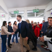 Foto de Leite usando blazer escuro e calça bege cumprimentando pessoas na entrada da escola. Há um grupo de pessoas sorrindo, e bandeirinhas de festa junina decoram o espaço.