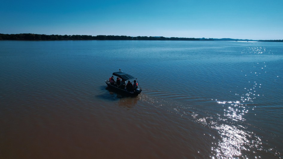 Uma vista aérea de um rio largo e calmo sob um céu azul claro. No centro, um pequeno barco com quatro pessoas a bordo navega da esquerda para a direita, deixando um leve rastro na água. A água do rio tem duas tonalidades distintas: a porção mais próxima da câmera é de um marrom-barrento, enquanto a parte mais distante é azul-escura, refletindo o céu. À direita, a luz do sol brilha intensamente na superfície da água. Na linha do horizonte, a vegetação ribeirinha é visível sob o céu azul claro e sem nuvens.