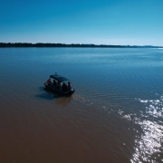 Uma vista aérea de um rio largo e calmo sob um céu azul claro. No centro, um pequeno barco com quatro pessoas a bordo navega da esquerda para a direita, deixando um leve rastro na água. A água do rio tem duas tonalidades distintas: a porção mais próxima da câmera é de um marrom-barrento, enquanto a parte mais distante é azul-escura, refletindo o céu. À direita, a luz do sol brilha intensamente na superfície da água. Na linha do horizonte, a vegetação ribeirinha é visível sob o céu azul claro e sem nuvens.