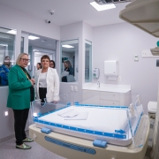 A imagem mostra uma sala de hospital, possivelmente uma unidade neonatal, com um berço aquecido em primeiro plano. Duas pessoas estão conversando ao lado do berço. Arita Bergmann está vestida com um blazer verde e a outra com um blazer branco. Ao fundo, há mais pessoas observando através de uma janela de vidro. A sala é bem iluminada e possui equipamentos médicos nas paredes e no teto.