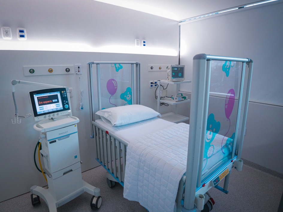 A imagem mostra um quarto de hospital com uma cama pediátrica. A cama tem grades laterais decoradas com imagens de ursinhos e balões. Há equipamentos médicos ao redor da cama, incluindo um ventilador à esquerda e um monitor à direita. O quarto é limpo, bem iluminado e parece ser projetado para cuidados pediátricos.