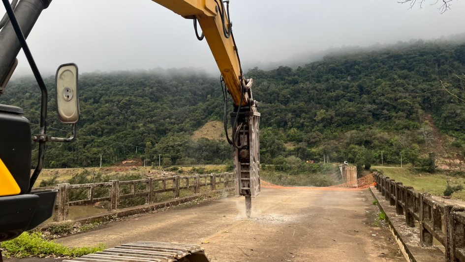 A imagem mostra uma escavadeira hidráulica amarela, com a marca "Ekiterra" visível no braço, operando na demolição da pista de uma ponte antiga. A máquina está com o martelete perfurando o concreto da ponte. A ponte, que parece ser de concreto e ferro, está visivelmente danificada. Ao fundo, há uma paisagem montanhosa densamente arborizada, com as montanhas parcialmente encobertas por névoa ou nuvens baixas. O tempo parece nublado.
