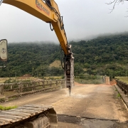 A imagem mostra uma escavadeira hidráulica amarela, com a marca "Ekiterra" visível no braço, operando na demolição da pista de uma ponte antiga. A máquina está com o martelete perfurando o concreto da ponte. A ponte, que parece ser de concreto e ferro, está visivelmente danificada. Ao fundo, há uma paisagem montanhosa densamente arborizada, com as montanhas parcialmente encobertas por névoa ou nuvens baixas. O tempo parece nublado.
