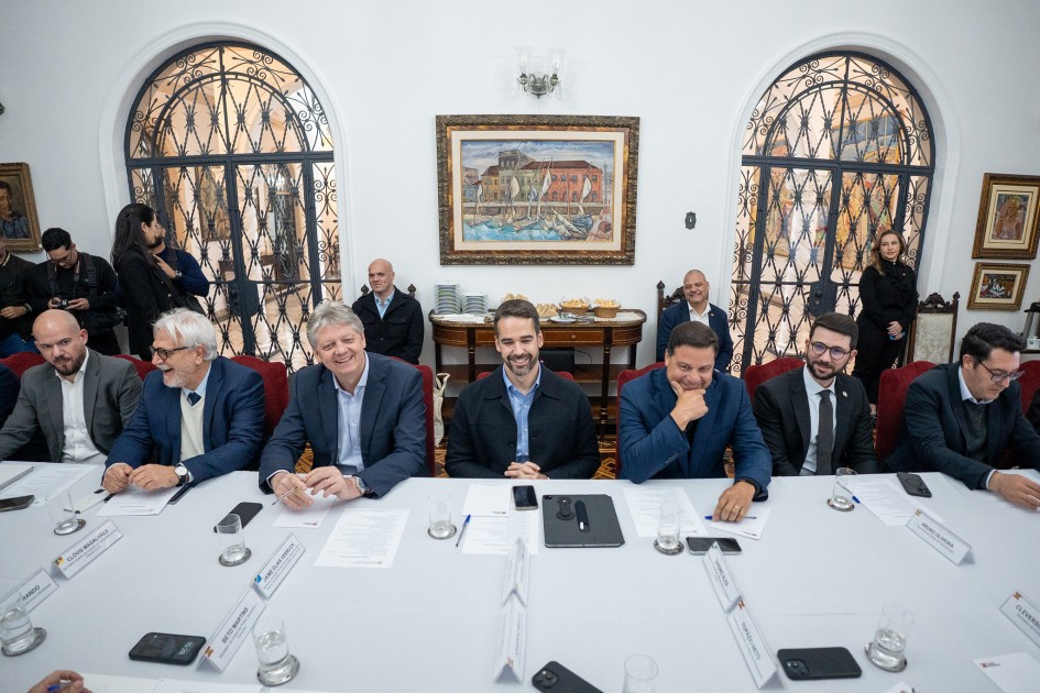 A imagem mostra um grupo de homens, a maioria de terno, sentados em volta de uma grande mesa de reunião oval branca. No centro da mesa, sorrindo e olhando para a frente, está o governador Eduardo Leite. Ao lado dele, à direita, está o governador do Paraná, Ratinho Júnior, e à esquerda, o governador de Santa Catarina, Jorginho Mello. Os participantes estão sentados com documentos, copos e celulares à sua frente. Ao fundo, a sala possui uma decoração clássica com janelas arqueadas e gradeadas, e um grande quadro pendurado na parede central. Há outras pessoas, em pé, observando a reunião.
