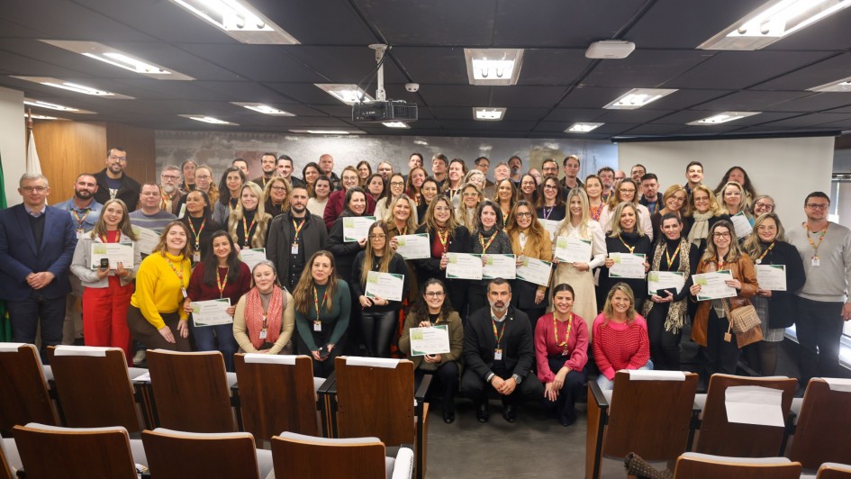 A foto, em ambiente fechado, mostra um grande grupo de pessoas, predominantemente adultos, posando para uma foto de formatura da turma da Academia de Líderes. A maioria está em pé na parte de trás, enquanto algumas pessoas, incluindo homens e mulheres, estão sentadas na frente, exibindo diplomas ou certificados. Muitos sorriem para a câmera. Ao fundo, uma parede escura e painéis de madeira são visíveis, junto a uma bandeira discreta à esquerda. A iluminação do teto é clara e uniforme.