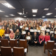 A foto, em ambiente fechado, mostra um grande grupo de pessoas, predominantemente adultos, posando para uma foto de formatura da turma da Academia de Líderes. A maioria está em pé na parte de trás, enquanto algumas pessoas, incluindo homens e mulheres, estão sentadas na frente, exibindo diplomas ou certificados. Muitos sorriem para a câmera. Ao fundo, uma parede escura e painéis de madeira são visíveis, junto a uma bandeira discreta à esquerda. A iluminação do teto é clara e uniforme.