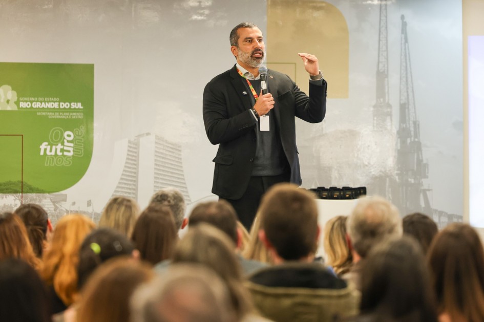 A foto mostra um evento em ambiente interno mostra o secretário-adjunto da SPGG, Bruno Silveira, no centro, em pé no palco, gesticulando com a mão esquerda enquanto fala a uma plateia. Ele é um homem de pele morena, barba e cabelo grisalhos, vestindo um terno escuro sobre um suéter cinza e uma camisa branca. O microfone está preso à sua lapela. Atrás dele, há um telão com a logo do Governo do Estado do Rio Grande do Sul e a frase "O futuro nos une" em verde e branco à esquerda. A plateia, composta por várias cabeças em primeiro plano e embaçada, está de costas para a câmera, olhando para o orador. A iluminação é clara e focada no palco.