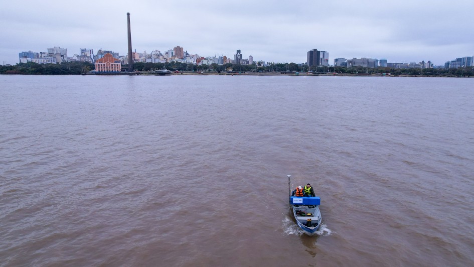 Uma vista aérea mostra um barco pequeno de pesquisa, cinza e azul, navegando em um rio de águas turvas sob um céu nublado. Duas pessoas, vestindo coletes salva-vidas amarelos, estão a bordo do barco, que se move da direita para o centro da imagem, deixando um pequeno rastro na água. No horizonte, a cidade de Porto Alegre se estende com uma silhueta de edifícios variados. Um dos pontos mais proeminentes é uma alta chaminé industrial escura no centro-esquerda da paisagem urbana. Prédios altos e a orla da cidade são visíveis sob o céu cinzento.