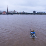 Uma vista aérea mostra um barco pequeno de pesquisa, cinza e azul, navegando em um rio de águas turvas sob um céu nublado. Duas pessoas, vestindo coletes salva-vidas amarelos, estão a bordo do barco, que se move da direita para o centro da imagem, deixando um pequeno rastro na água. No horizonte, a cidade de Porto Alegre se estende com uma silhueta de edifícios variados. Um dos pontos mais proeminentes é uma alta chaminé industrial escura no centro-esquerda da paisagem urbana. Prédios altos e a orla da cidade são visíveis sob o céu cinzento.