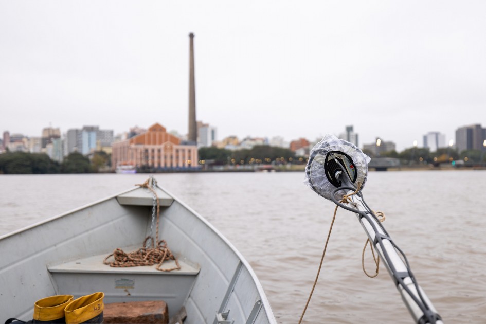 A fotografia, tirada da proa de um pequeno barco de metal cinza, mostra equipamentos de medição estendendo-se sobre a água turva do rio. Uma corda enrolada e um par de botas de borracha amarelas estão visíveis no interior do barco, em primeiro plano. O equipamento de medição, parcialmente coberto por um plástico transparente, está posicionado no lado direito da imagem. Ao fundo, a silhueta da cidade de Porto Alegre se eleva sob um céu nublado. Destacam-se edifícios variados, incluindo uma estrutura proeminente com telhado de terracota, e uma alta chaminé industrial escura dominando o horizonte. A água do rio é cinzenta e calma, refletindo a atmosfera do dia nublado.