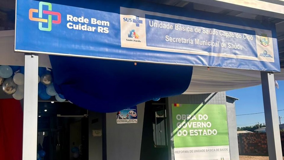 A foto exibe a fachada da Unidade Básica de Saúde em Capão do Cipó, reformada e pertencente à Rede Bem Cuidar e à Estratégia de Saúde da Família. Acima da entrada, uma grande faixa azul e branca se estende. O lado esquerdo da faixa apresenta um logotipo verde e azul com uma cruz e um coração, e o texto "Rede Bem Cuidar RS". O lado direito da faixa exibe o logotipo do SUS e "Unidade Básica de Saúde Capão do Cipó", seguido por "Secretaria Municipal de Saúde". Abaixo da faixa, à direita da entrada, há um painel verde com os dizeres "OBRA DO GOVERNO DO ESTADO" em letras brancas, e "REFORMA DE UNIDADE BÁSICA DE SAÚDE". À esquerda da entrada, balões azuis e brancos adornam o local, e um pequeno cartaz é visível em uma parede. A construção é de cor clara, com detalhes em azul. O dia está ensolarado.