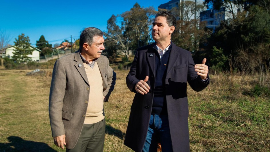 Foto dos dois homens estão de pé, conversando em um terreno aberto com grama seca. Adiló é mais velho, veste blazer bege e calça verde-oliva. Fabricio é mais jovem, usa sobretudo escuro sobre uma camisa branca e suéter azul-marinho. Ao fundo, há árvores, algumas casas e prédios. O céu está claro e azul.