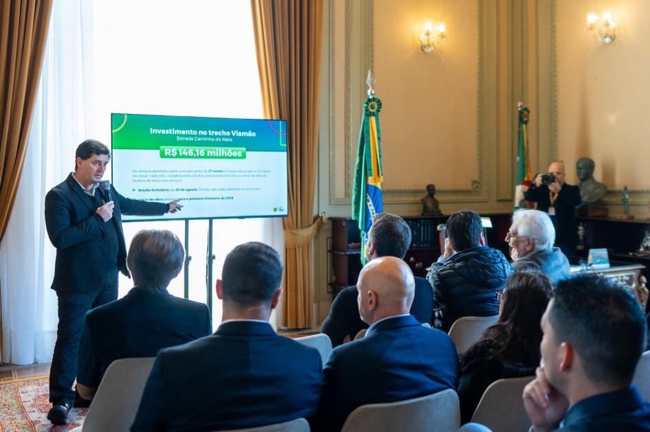 Caumo, de de terno escuro, aponta para a tela da TV, que exibe o valor “R$ 146,16 milhões”. À frente dele, várias pessoas estão sentadas assistindo. Ao fundo, cortinas douradas, bandeiras e móveis antigos.