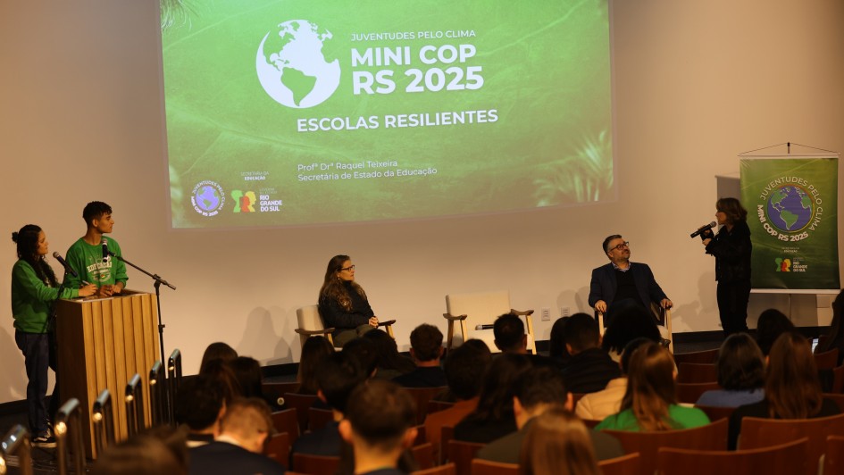 A foto mostra um evento sendo realizado em um auditório. No palco, à esquerda, dois jovens estudantes, um homem e uma mulher, ambos vestindo camisetas verdes, estão de pé ao lado de um púlpito de madeira claro. O homem está falando ao microfone. No centro do palco, duas pessoas sentam em poltronas brancas, uma mulher de óculos e um homem de blazer escuro, ambos olhando para frente. Mais à direita, outra mulher de pé, vestindo preto, segura um microfone e interage com o público ou com alguém fora do quadro. No fundo, um grande telão projeta um slide verde com a escrita "JUVENTUDES PELO CLIMA MINI COP RS 2025" e "ESCOLAS RESILIENTES", junto a logos do "GOVERNO DO ESTADO RIO GRANDE DO SUL" e "SECRETARIA DE ESTADO DA EDUCAÇÃO". À direita do palco, um banner verde com a mesma temática está visível. As primeiras filas do auditório estão cheias de estudantes, visíveis da parte de trás da cabeça, sentados em cadeiras de madeira. A iluminação é focada no palco.