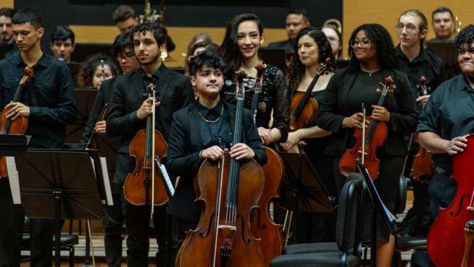A foto mostra um palco com vários músicos da Ospa Jovem, predominantemente jovens e adultos, tocando instrumentos de corda. Em primeiro plano, no centro, uma pessoa de cabelo escuro e camisa preta segura um violoncelo, olhando para o lado. À sua direita, uma mulher sorri enquanto segura um violino, vestindo uma blusa preta brilhante. Outros músicos ao redor seguram violinos e violoncelos, alguns olhando para frente e outros para os lados. Ao fundo, músicos da orquestra sentam em fileiras, com instrumentos de sopro e percussão visíveis, todos vestindo preto. O palco possui um fundo de madeira lisa e uma iluminação amarelada que destaca os instrumentistas.