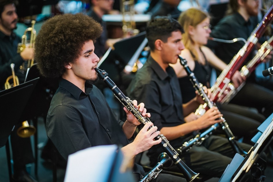A foto mostra um jovem músico da Ospa Jovem, de pele escura e cabelo crespo, tocando clarinete em primeiro plano, sentado em uma cadeira. Ele veste uma camisa preta e está concentrado no instrumento. Atrás dele, ligeiramente desfocado, outro músico também toca clarinete, enquanto à direita, um terceiro músico toca um fagote. Ao fundo, outras partes da orquestra são visíveis, incluindo instrumentos de sopro e cordas. A iluminação é focada nos músicos, criando um contraste com o fundo mais escuro do palco.