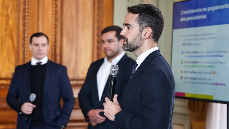 A imagem mostra três homens vestindo ternos, cada um com um microfone em mãos, como se estivessem apresentando ou debatendo algo em público. Eles estão em um ambiente fechado que se assemelha a uma sala de conferência ou reunião formal. Quem está com o microfone é o governador Eduardo Leite. Ao fundo, há um grande painel com informações sobre o pagamento de precatórios - que são dívidas judiciais do governo.