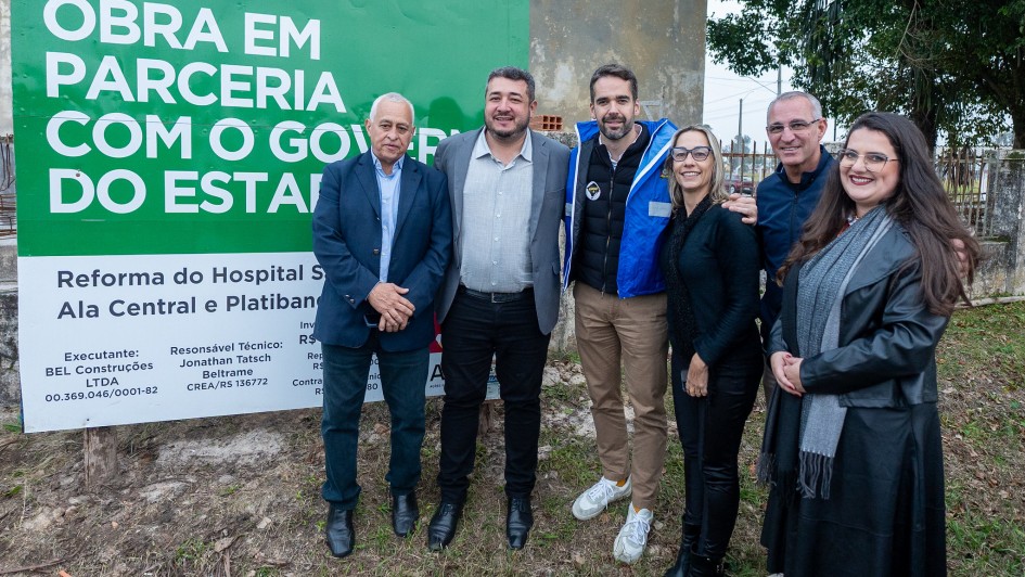 A imagem mostra um grupo de seis pessoas, quatro homens e duas mulheres, em pé ao ar livre, em frente a um grande banner verde. O banner exibe em letras brancas a frase "OBRA EM PARCERIA COM O GOVERNO DO ESTADO" e informações sobre uma "Reforma do Hospital", incluindo o nome da construtora "BEL Construções LTDA". As pessoas estão sorrindo e olhando para a câmera. O governador Eduardo Leite está presente, vestindo uma jaqueta azul sobre um moletom. À esquerda do governador, um homem de terno. À direita, uma mulher de preto com um cachecol e outro homem. Ao fundo, há árvores e uma construção.