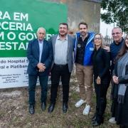 A imagem mostra um grupo de seis pessoas, quatro homens e duas mulheres, em pé ao ar livre, em frente a um grande banner verde. O banner exibe em letras brancas a frase "OBRA EM PARCERIA COM O GOVERNO DO ESTADO" e informações sobre uma "Reforma do Hospital", incluindo o nome da construtora "BEL Construções LTDA". As pessoas estão sorrindo e olhando para a câmera. O governador Eduardo Leite está presente, vestindo uma jaqueta azul sobre um moletom. À esquerda do governador, um homem de terno. À direita, uma mulher de preto com um cachecol e outro homem. Ao fundo, há árvores e uma construção.