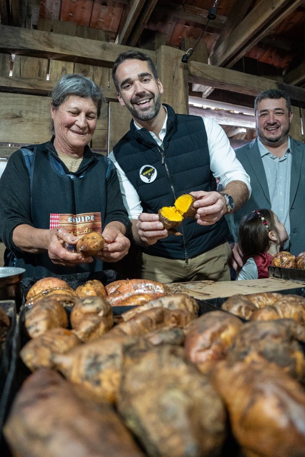 A imagem mostra o governador Eduardo Leite no centro, sorrindo e segurando uma batata-doce cortada ao meio, mostrando seu interior alaranjado. Ele está vestindo uma camisa branca e um colete escuro. À esquerda, uma mulher com um avental e sorriso no rosto segura uma batata-doce inteira. À direita, atrás do governador, outro homem sorri para a câmera, e uma criança pequena é parcialmente visível abaixo dele. Em primeiro plano, há uma grande quantidade de batatas-doces assadas ou cozidas sobre uma superfície escura. O ambiente parece ser rústico, com estruturas de madeira visíveis ao fundo, sugerindo uma feira ou evento rural.