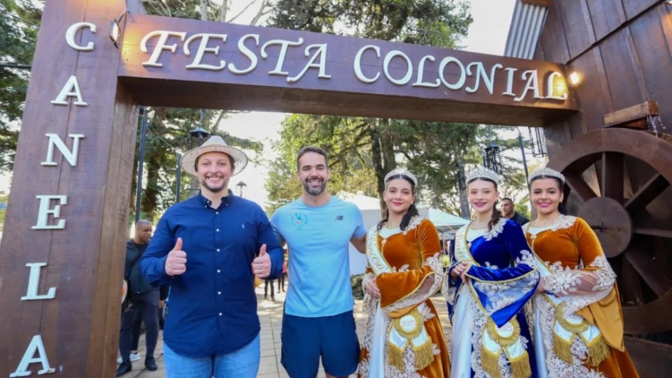 A fotografia mostra cinco pessoas (o governador Eduado Leite, o prefeito Gilberto Cezar e três mulheres da corte, posando sob um arco de madeira decorativo. Esse arco tem palavras em destaque: no topo está escrito "Festa Colonial" e na lateral esquerda, "Canela", indicando que o local é uma festa tradicional na cidade de Canela.