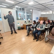 A foto mostra uma sala de treinamento ou aula com um grupo de aproximadamente vinte pessoas. Um homem com barba e camisa escura está em pé à esquerda, gesticulando e aparentemente conduzindo uma apresentação ou palestra. À sua esquerda, há uma tela grande montada em um suporte, mostrando algo fora de foco. A plateia está sentada em cadeiras de escritório, olhando para o apresentador. As pessoas são de diversas idades e vestem roupas casuais. A sala tem paredes claras com detalhes em madeira, um chão de laminado claro e iluminação fluorescente no teto. Há também janelas que parecem dar para outros espaços de escritório. Algumas mesas e objetos como garrafas de água estão visíveis na sala. O ambiente sugere um aprendizado ou uma reunião de trabalho.