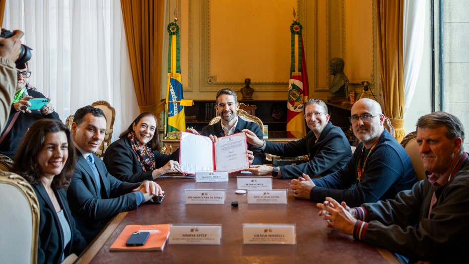 Uma mesa retangular de madeira em uma sala com cortinas douradas, duas bandeiras (Brasil e Rio Grande do Sul) e iluminação clássica ao fundo. O governador Eduardo Leite está ao centro, sorrindo e segurando com outra pessoa um documento aberto. Ao redor, sete pessoas sentadas, também sorrindo. Placas com nomes identificam alguns participantes.