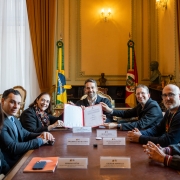 Uma mesa retangular de madeira em uma sala com cortinas douradas, duas bandeiras (Brasil e Rio Grande do Sul) e iluminação clássica ao fundo. O governador Eduardo Leite está ao centro, sorrindo e segurando com outra pessoa um documento aberto. Ao redor, sete pessoas sentadas, também sorrindo. Placas com nomes identificam alguns participantes.