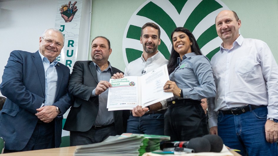 Esta é uma fotografia colorida de cinco pessoas adultas, quatro homens e uma mulher, posando lado a lado em um evento formal de assinatura de documentos.
Da esquerda para a direita:
Homem idoso, de óculos: Veste um terno azul-marinho com camisa branca e gravata escura. Ele sorri para a câmera.
Homem de meia-idade: Veste um terno escuro com camisa clara. Ele está segurando uma folha de papel branca com as duas mãos, que parece ser um documento ou um certificado, e olha para a frente.
Homem de meia-idade no centro: Veste uma camisa social clara de mangas compridas e calça escura. Ele também segura o mesmo documento que o homem à sua esquerda e sorri para a câmera. Há um logotipo com a frase "Jovens Pelo Futuro da Agricultura Rural" no documento.
Mulher jovem: Veste uma camisa cinza-claro de mangas compridas e calça escura. Ela sorri amplamente e segura a outra extremidade do documento, que parece estar aberto em uma pasta ou caderno.
Homem idoso, à direita: Veste uma camisa social branca de mangas compridas e calça jeans. Ele sorri e olha para a câmera.
Ao fundo, há uma parede verde clara com um grande logotipo estilizado em verde escuro, que se assemelha a um círculo com elementos pontiagudos. Há também um banner vertical com a letra "JUR" e uma mão estilizada segurando algo.
Na frente dos indivíduos, sobre uma mesa de madeira escura, há uma pilha de papéis, alguns cadernos e um microfone preto, sugerindo que se trata de uma coletiva de imprensa ou um evento de assinatura. A iluminação é boa e a imagem é nítida.