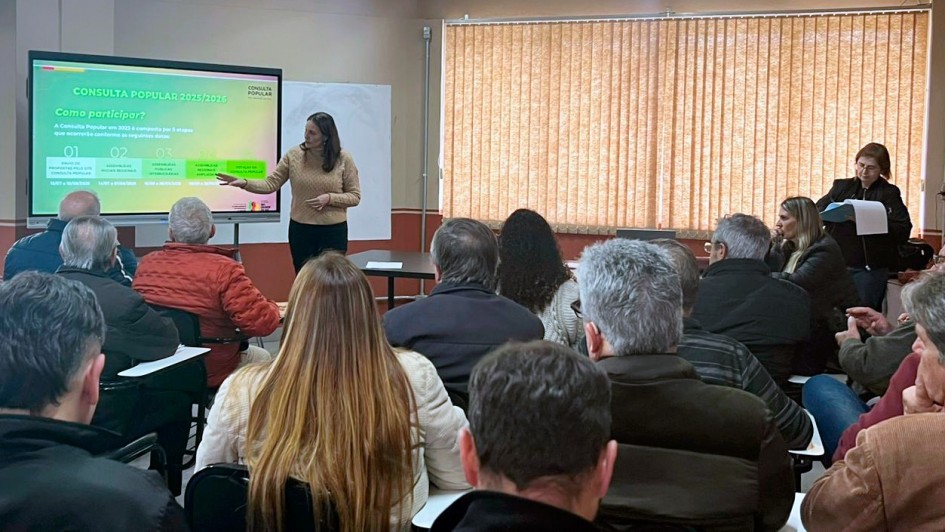 Esta é uma fotografia tirada de um ângulo posterior da plateia, mostrando uma apresentação em uma sala de aula ou auditório pequeno.
No centro da imagem, em primeiro plano, várias pessoas estão sentadas de costas para o observador, olhando para frente, onde uma mulher está apresentando. A plateia é composta principalmente por adultos e idosos.
À esquerda, no fundo, uma mulher vestindo uma blusa bege clara está em pé ao lado de uma grande tela de projeção branca. Na tela, há um slide com o título "CONSULTA POPULAR 2025/2026" e "Como participar?", com informações divididas em três etapas numeradas (01, 02, 03) e caixas de texto abaixo. A mulher aponta para a tela com a mão direita.
À direita, no fundo da sala, outra mulher está em pé, atrás de uma mesa, com alguns papéis na mão. A sala tem paredes claras e uma grande janela coberta por persianas verticais bege no lado direito. O teto é de cor clara, com luminárias embutidas. A iluminação é boa e a imagem é nítida.