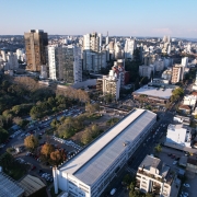 A foto mostra uma vista aérea panorâmica da cidade de Caxias do Sul durante o dia, sob um céu claro e azul. Em primeiro plano, no centro da imagem, destaca-se um longo edifício retangular com telhado claro, cercado por uma área de estacionamento com muitos carros e uma área verde arborizada à esquerda. Ao redor, a paisagem é dominada por uma densidade de edifícios de diferentes alturas e estilos, que se estendem até o horizonte. Há tanto prédios mais antigos quanto construções modernas de múltiplos andares. No canto inferior direito, uma rua movimentada com carros visíveis, sugerindo a vida urbana da cidade. A iluminação solar suave indica um fim de tarde ou começo de manhã, realçando os contornos dos edifícios e a vasta extensão urbana.