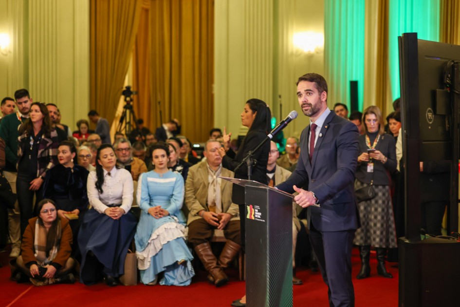 Esta é uma fotografia de um evento público em um salão opulento, onde um homem está discursando no primeiro plano à direita.
O homem, com barba, veste um terno azul-marinho, camisa branca e gravata, e está de pé atrás de um púlpito escuro com o logotipo "RS". Ele segura um microfone com a mão direita e gesticula com a esquerda enquanto fala.
Atrás dele e à esquerda, uma grande plateia está sentada em cadeiras sobre um tapete vermelho. Algumas pessoas na plateia vestem trajes típicos gaúchos: duas mulheres sentadas na frente vestem vestidos longos azuis e brancos com babados, e um homem sentado ao lado delas veste bombachas e camisa tradicional. Uma mulher de cabelos longos e escuros está em pé atrás do púlpito, olhando para o orador.
O salão possui paredes em tom creme com colunas decoradas e grandes cortinas douradas ao fundo. Há uma iluminação verde e branca vinda do fundo. O ambiente é grandioso e sugere um evento cultural ou oficial.