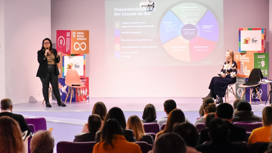 A imagem retrata uma apresentação em um auditório, com duas mulheres no palco e uma plateia sentada de costas para o observador.

No centro do palco, à esquerda, uma mulher de óculos, blusa com estampa de onça e calças escuras está em pé, segurando um microfone e gesticulando enquanto fala. Atrás dela, há um totem com o logotipo dos 17 Objetivos de Desenvolvimento Sustentável (ODS) da ONU, destacando os objetivos "10 Redução das Desigualdades" e "12 Consumo e Produção Responsáveis".

À direita, sentada em uma cadeira, está outra mulher, vestindo uma blusa estampada de flores e uma saia escura.

No fundo do palco, um grande telão exibe um infográfico circular colorido, com o título "Descarbonização Rio Grande do Sul". O gráfico apresenta seções como "Inovação", "Ambiente de Negócios", "Descarbonização", "Infraestrutura", "Gestão Urbana", "Governo Climático" e "Educação". Além disso, o telão lista tópicos como "Transição Energética Justa", "Pagamento por Serviços Ambientais", "Agenda Internacional", "Monitoramento da Qualidade do Ar", "Educação Climática e Justiça Ambiental", "Governança Climática" e "Inovação".

À direita do telão, há um banner com o título "11º Fórum Gaúcho de Mudanças Climáticas", acompanhado de outros logotipos relacionados aos ODS. A plateia ocupa as primeiras filas do auditório, com cadeiras roxas, e a maioria das pessoas está de costas, prestando atenção à apresentação.