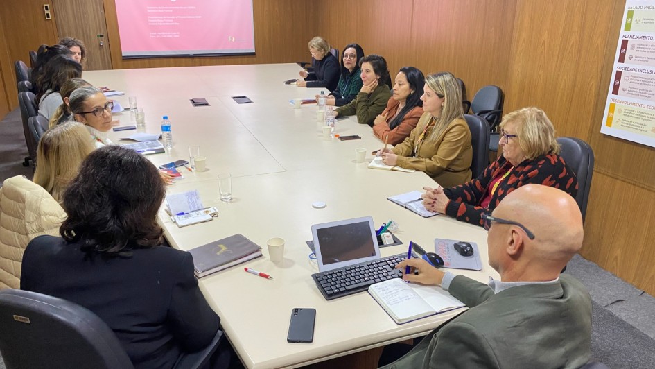 A imagem mostra uma sala de reunião com um grupo de pessoas sentadas ao redor de uma grande mesa retangular, que é bege clara. A maioria das pessoas são mulheres, com idades variadas, e todas parecem atentas, algumas olhando para o centro da mesa ou para a tela de projeção.
No primeiro plano, de costas para o observador, um homem calvo com óculos veste uma jaqueta verde oliva e está usando um notebook e anotando algo em um caderno. Ao lado dele, há um smartphone preto.
No fundo da sala, um projetor está ligado e exibe a palavra "Obrigada!" em letras brancas sobre um fundo rosa claro na parede. Há também um banner vertical parcialmente visível à direita, com a palavra "MAPA" no topo.
A mesa está repleta de papéis, cadernos, copos com água e alguns laptops, indicando uma reunião de trabalho. As cadeiras são escuras, com braços, e as paredes da sala são de um tom claro, com painéis de madeira.