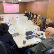 A imagem mostra uma sala de reunião com um grupo de pessoas sentadas ao redor de uma grande mesa retangular, que é bege clara. A maioria das pessoas são mulheres, com idades variadas, e todas parecem atentas, algumas olhando para o centro da mesa ou para a tela de projeção.

No primeiro plano, de costas para o observador, um homem calvo com óculos veste uma jaqueta verde oliva e está usando um notebook e anotando algo em um caderno. Ao lado dele, há um smartphone preto.

No fundo da sala, um projetor está ligado e exibe a palavra "Obrigada!" em letras brancas sobre um fundo rosa claro na parede. Há também um banner vertical parcialmente visível à direita, com a palavra "MAPA" no topo.

A mesa está repleta de papéis, cadernos, copos com água e alguns laptops, indicando uma reunião de trabalho. As cadeiras são escuras, com braços, e as paredes da sala são de um tom claro, com painéis de madeira.