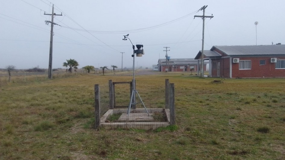A imagem mostra uma estação meteorológica automática instalada em uma área gramada, cercada por uma pequena estrutura de madeira com tela metálica. O equipamento está montado sobre um tripé metálico, com sensores no topo. Ao fundo, há postes de energia, algumas palmeiras e prédios de alvenaria com telhado de duas águas, ar-condicionado e janelas fechadas. O céu está nublado ou com névoa, indicando um clima úmido ou frio