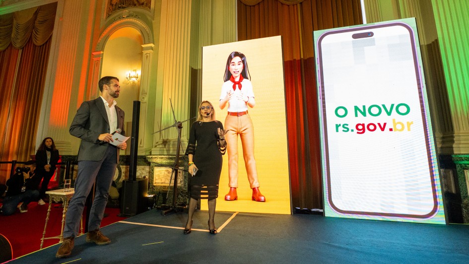Um homem e uma mulher estão em um palco: eles egura um microfone, ela, um celular. Ao fundo, há um painel com um desenho animado de mulher em roupa social. Ao lado, outro painel exibe um celular e a frase “O NOVO rs.gov.br”. O ambiente é de uma apresentação oficial.