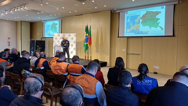 A foto mostra um seminário internacional da Defesa Civil ocorrendo em um auditório moderno. No centro do palco, um orador de costas para o observador, vestido com colete da Defesa Civil, fala para uma plateia atenta, composta em sua maioria por homens e mulheres também usando coletes laranjas ou uniformes escuros. Atrás do orador, em destaque, há duas bandeiras: uma do Brasil e outra do Rio Grande do Sul. Duas grandes telas de projeção, uma em cada lado do palco, exibem mapas coloridos da Europa, com destaque para a Itália, e outros dados relacionados. A iluminação é clara e uniforme, e o ambiente é organizado, com fileiras de cadeiras preenchidas. Alguns participantes usam fones de ouvido para tradução simultânea. A imagem sugere um evento de cooperação e aprendizado sobre temas de proteção civil.