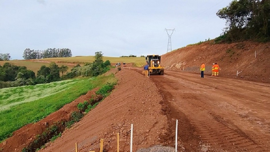 A imagem mostra uma estrada de chão em obras, com uma máquina ao centro e alguns trabalhadores uniformizados na pista. 