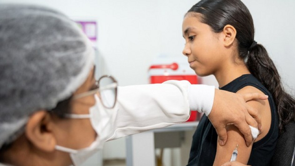 A foto mostra uma jovem de pele morena clara, com cabelos pretos longos presos em um rabo de cavalo lateral, sentada de perfil, recebendo uma vacina no braço esquerdo. Ela usa uma camiseta preta e observa a agulha com uma expressão séria e atenta. A agulha, fina e prateada, é visível no momento da aplicação, com o braço dela levemente flexionado. À frente da jovem, uma profissional de saúde, parcialmente visível, com touca branca, máscara branca e óculos, está aplicando a injeção. A mão da profissional segura o braço da jovem suavemente, e um chumaço de algodão branco é visível próximo ao ponto da aplicação. O ambiente ao fundo é um local de saúde, com paredes brancas, e alguns elementos médicos, como caixas de descarte de material ou kits de primeiros socorros em tons de vermelho e branco, ligeiramente desfocados.