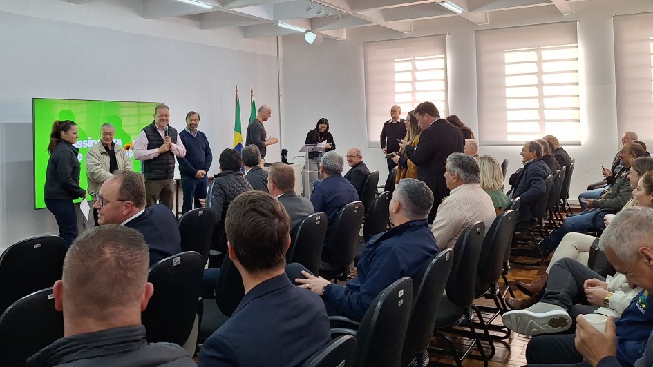 Uma foto mostra uma reuni&atilde;o em ambiente fechado com muitas pessoas, algumas em p&eacute; e outras sentadas. &Agrave; esquerda, v&aacute;rios homens est&atilde;o de p&eacute; perto de uma grande tela verde brilhante com algum texto vis&iacute;vel. Uma mulher est&aacute; &agrave; extrema esquerda deles, olhando para eles. No centro e &agrave; direita, uma plateia est&aacute; sentada em fileiras de cadeiras escuras, de frente para a parte da frente onde est&atilde;o os indiv&iacute;duos em p&eacute;. Alguns membros da plateia est&atilde;o olhando para a frente, enquanto outros est&atilde;o olhando para seus telefones ou conversando entre si. A sala tem paredes de cores claras e janelas com persianas s&atilde;o vis&iacute;veis no lado direito. A ilumina&ccedil;&atilde;o geral &eacute; clara.